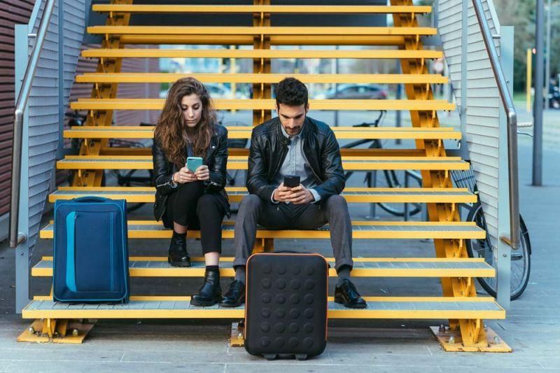 Genrebild över en man och en kvinna som väntar med sina väskor sittande på en trappa med mobilen i handen