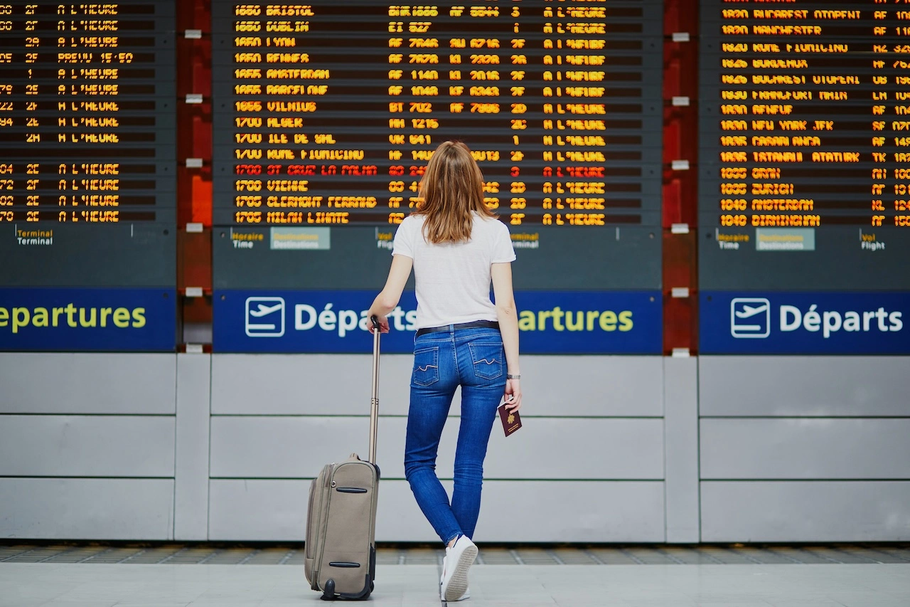 Mujer joven en un aeropuerto internacional con su equipaje y pasaporte cerca de la pantalla de informaci&oacute;n de vuelos