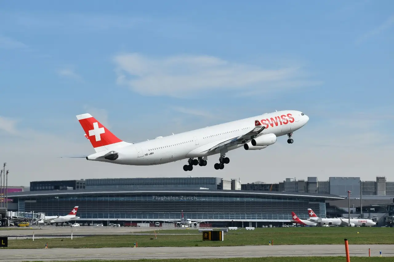 A red and white SWISS plane taking off with the Zurich Airport terminal below